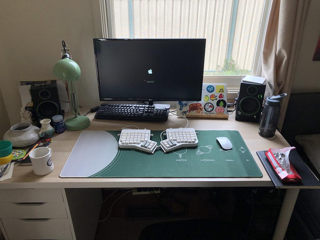 A desk in front of a window. There's a laptop in clamshell mode, a split keyboard and magic mouse among a bunch of other desk-related bits and pieces