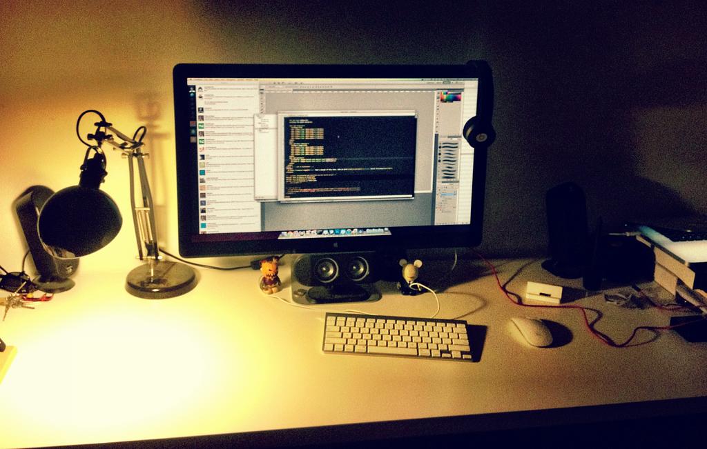 A dimly lit monitor with a magic mouse and keyboard in-front. The light-source is an IKEA lamp