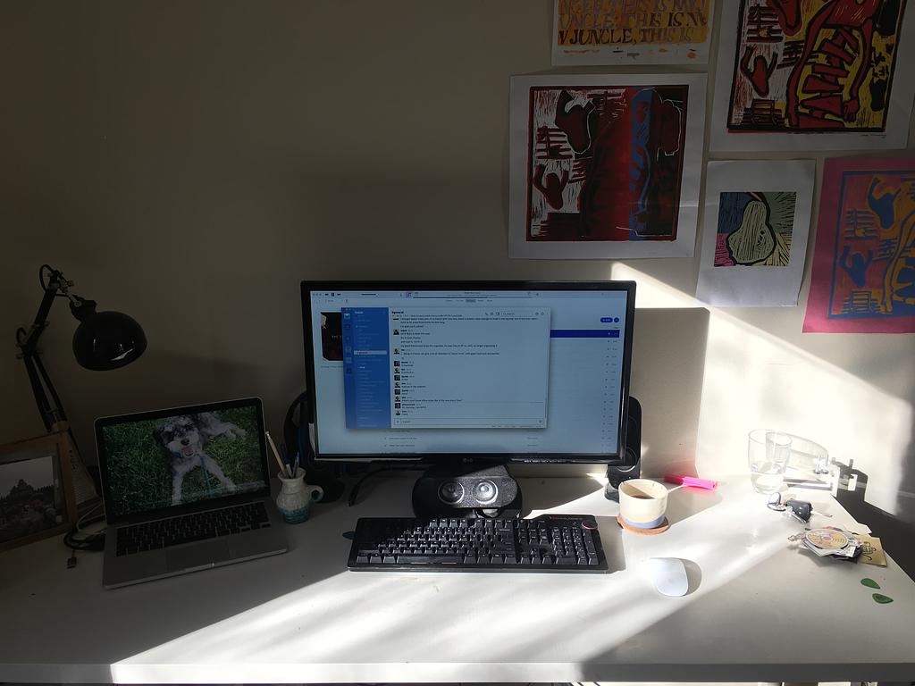 A desk lit by a beam of sunlight. On it is a laptop plugged in to a monitor, a magic mouse, a mechanical keyboard and a short coffee mug
