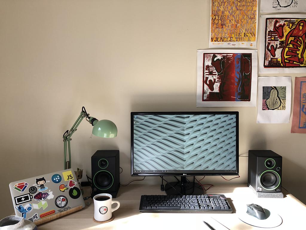 A desk with a laptop in clam-shell mode, a set of bookshelf speakers, a green lamp and a mechanical keyboard