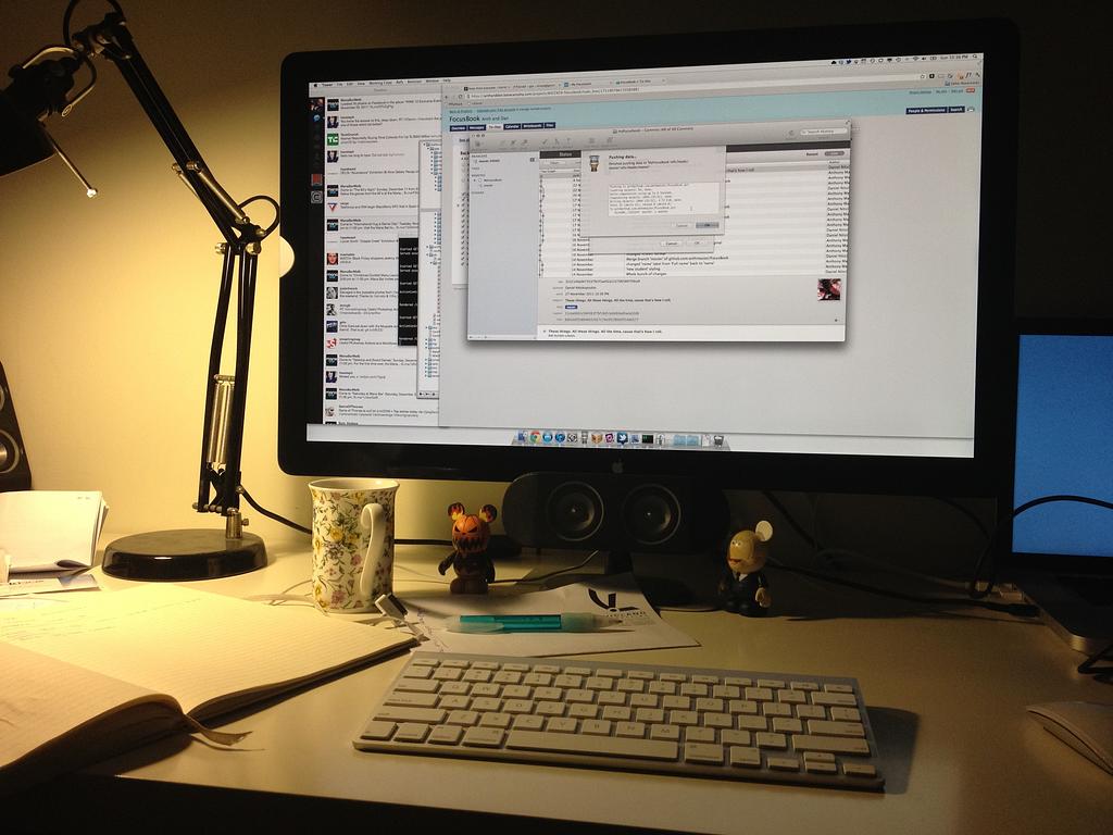 A dimly lit monitor with a magic keyboard, mug and notebook in front