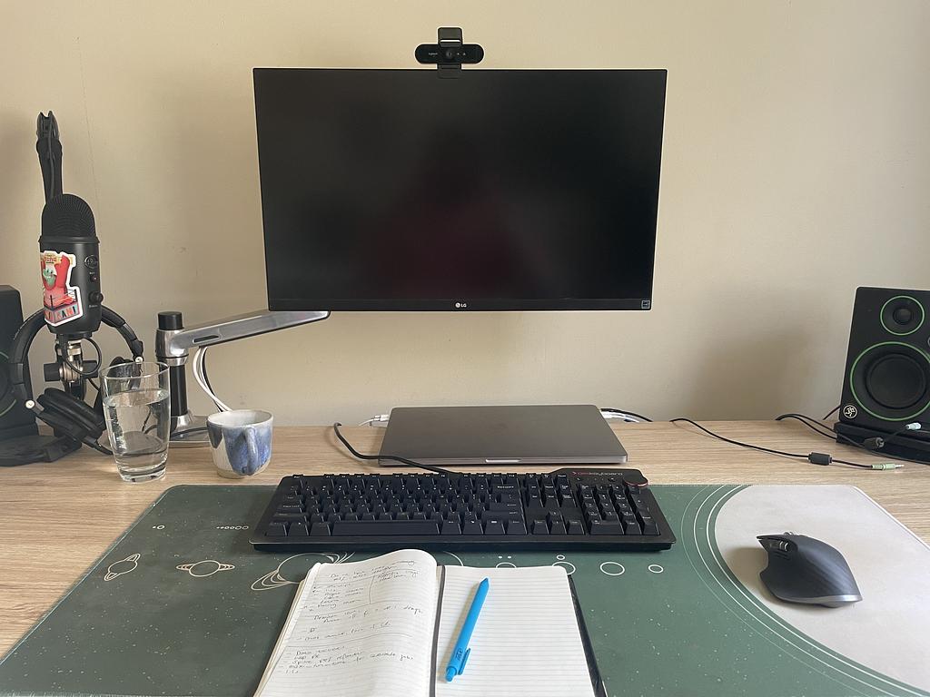 A desk with a monitor on an arm and raised well above the top of the desk. Also, a microphone, bookshelf speakers and a mechanical keyboard are in view